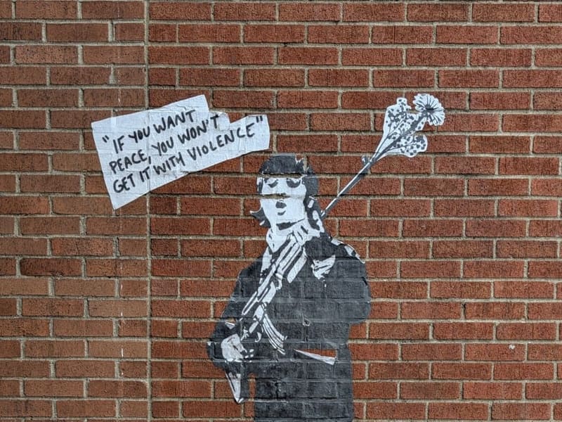 Stencil street art on brick: a figure with a flower in a gun barrel, 'If you want peace, you won't get it with violence'