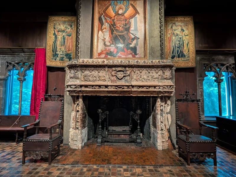 A carved stone fireplace beneath a religious painting in a dim Gardner Museum room