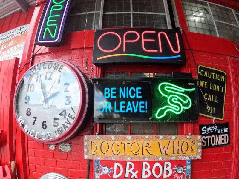 Glowing neon signs at Dr. Bob's Bywater folk-art studio: 'Open', 'Be Nice or Leave', 'Dr. Bob'