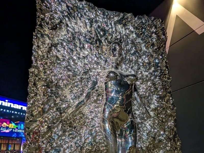 A polished chrome relief sculpture of a figure emerging from textured metal, lit at night