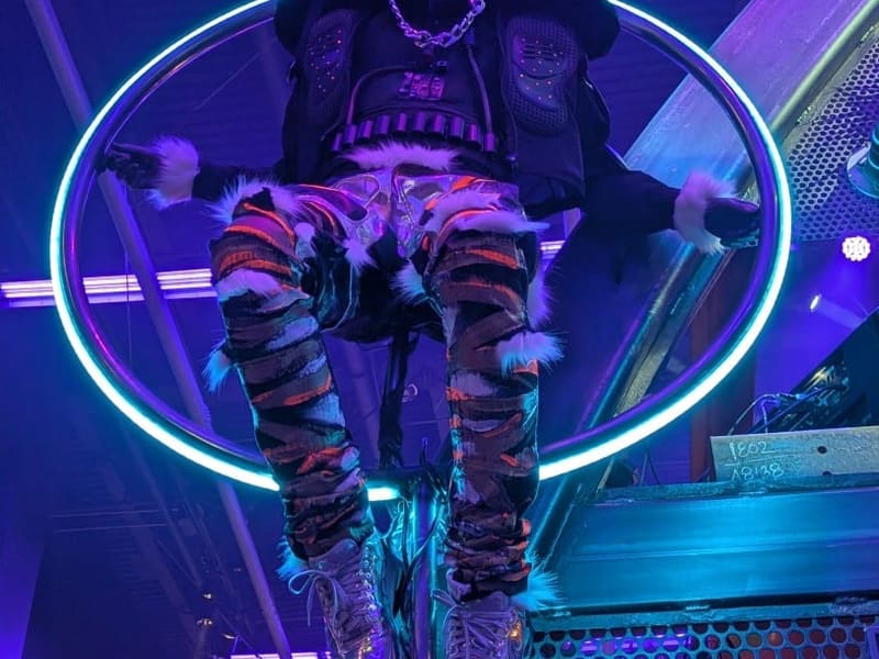 A neon-lit performer in a glowing tiger costume seated inside an illuminated hoop