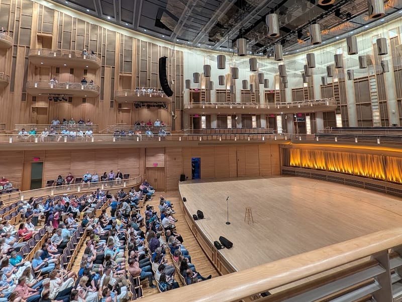 The wood-paneled Music Center concert hall with the audience seated before an empty stage and a lone microphone.