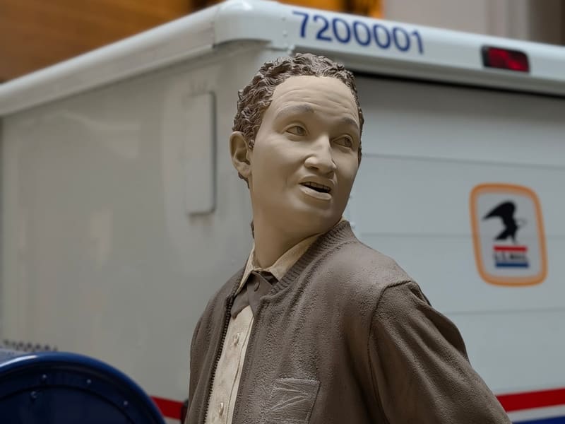 A lifelike figure of a modern USPS letter carrier beside a mail truck