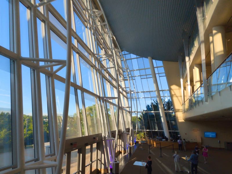 Interior lobby atrium at Strathmore: a tall curving glass wall with white steel bracing, a staircase in the foreground, and patrons gathered near the bar.
